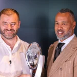FootballR - ELF - Zwei Männer posieren vor einem dunklen Hintergrund und lächeln beide. Der Mann links hält eine glänzende silberne Trophäe mit einer reflektierenden Oberfläche in Form eines Rugbyballs, die auf einem Ständer steht – ein Symbol ihres Sieges im Championship Game. Der Mann rechts trägt einen hellbraunen Nadelstreifenanzug, ein weißes Hemd und eine dunkle Krawatte. Diese Beschreibung wurde automatisch generiert.
