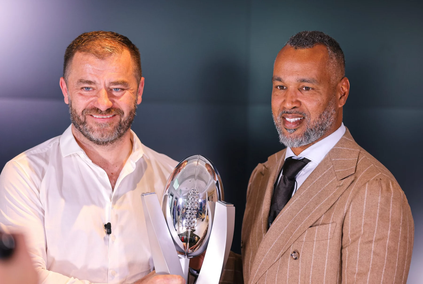FootballR - ELF - Zwei Männer posieren vor einem dunklen Hintergrund und lächeln beide. Der Mann links hält eine glänzende silberne Trophäe mit einer reflektierenden Oberfläche in Form eines Rugbyballs, die auf einem Ständer steht – ein Symbol ihres Sieges im Championship Game. Der Mann rechts trägt einen hellbraunen Nadelstreifenanzug, ein weißes Hemd und eine dunkle Krawatte. Diese Beschreibung wurde automatisch generiert.