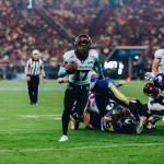 FootballR - NFL - Ein American-Football-Spieler mit kastanienbraunem Helm und weißem Trikot mit der Nummer 17 hält den Ball und läuft über das Feld. Im Hintergrund tackeln Spieler in Gelb, Blau, Kastanienbraun und Weiß. Ein Schiedsrichter in Streifen steht hinter ihnen, während sich das Stadion mit Zuschauern füllt. Es erinnert an ein Rhein Fire Spiel. Diese Beschreibung wurde automatisch generiert.