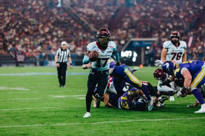 FootballR - NFL - Ein American-Football-Spieler mit kastanienbraunem Helm und weißem Trikot mit der Nummer 17 hält den Ball und läuft über das Feld. Im Hintergrund tackeln Spieler in Gelb, Blau, Kastanienbraun und Weiß. Ein Schiedsrichter in Streifen steht hinter ihnen, während sich das Stadion mit Zuschauern füllt. Es erinnert an ein Rhein Fire Spiel. Diese Beschreibung wurde automatisch generiert.