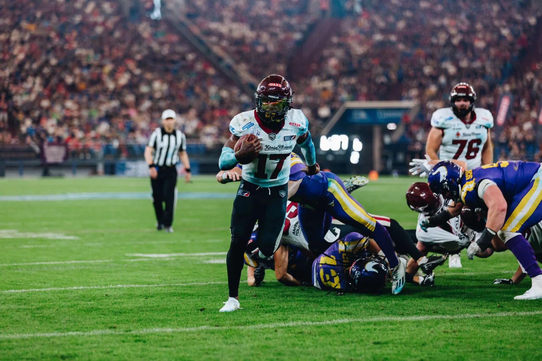 FootballR - NFL - Ein American-Football-Spieler mit kastanienbraunem Helm und weißem Trikot mit der Nummer 17 hält den Ball und läuft über das Feld. Im Hintergrund tackeln Spieler in Gelb, Blau, Kastanienbraun und Weiß. Ein Schiedsrichter in Streifen steht hinter ihnen, während sich das Stadion mit Zuschauern füllt. Es erinnert an ein Rhein Fire Spiel. Diese Beschreibung wurde automatisch generiert.