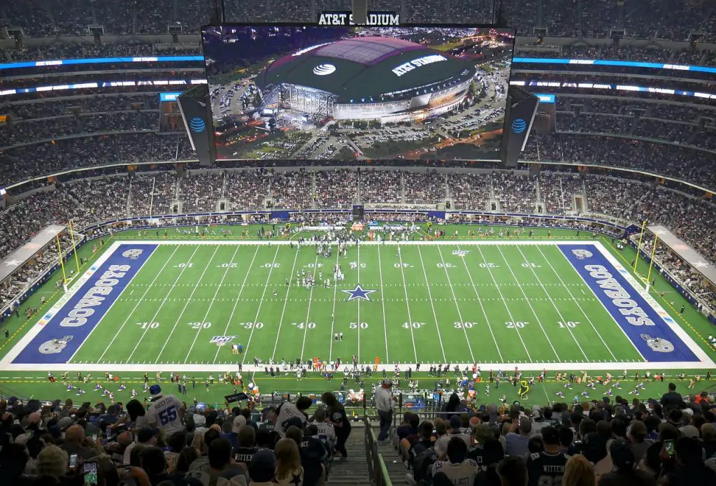 FootballR - NFL - Eine Weitwinkelansicht des AT&T-Stadions während eines Footballspiels der Dallas Cowboys. Das Spielfeld, umgeben von einer großen Zuschauermenge, zeigt das Sternenlogo der Cowboys an der 50-Yard-Linie und in jeder Endzone steht „Cowboys“. Während die kommende Saison näher rückt, machen die Fans aufgeregt NFL-Wetten, während sie das Spiel auf einem großen Bildschirm an der Decke verfolgen. Diese Beschreibung wurde automatisch generiert.