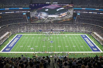 Markquese Bell - NFL - Klayton Adams - Eine Weitwinkelansicht des AT&T Stadium während eines Footballspiels der Dallas Cowboys. Das Spielfeld, umgeben von einer großen Zuschauermenge, zeigt das Sternenlogo der Cowboys an der 50-Yard-Linie und in jeder Endzone steht „Cowboys“. Während die kommende Saison näher rückt, machen die Fans aufgeregt NFL-Wetten, während sie das Spiel auf einem großen Bildschirm an der Decke verfolgen. Diese Beschreibung wurde automatisch generiert.