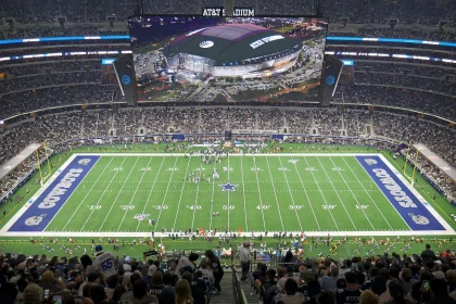 FootballR - NFL - Klayton Adams - Eine Weitwinkelansicht des AT&T Stadium während eines Footballspiels der Dallas Cowboys. Das Spielfeld, umgeben von einer großen Zuschauermenge, zeigt das Sternenlogo der Cowboys an der 50-Yard-Linie und in jeder Endzone steht „Cowboys“. Während die kommende Saison näher rückt, machen die Fans aufgeregt NFL-Wetten, während sie das Spiel auf einem großen Bildschirm an der Decke verfolgen. Diese Beschreibung wurde automatisch generiert.
