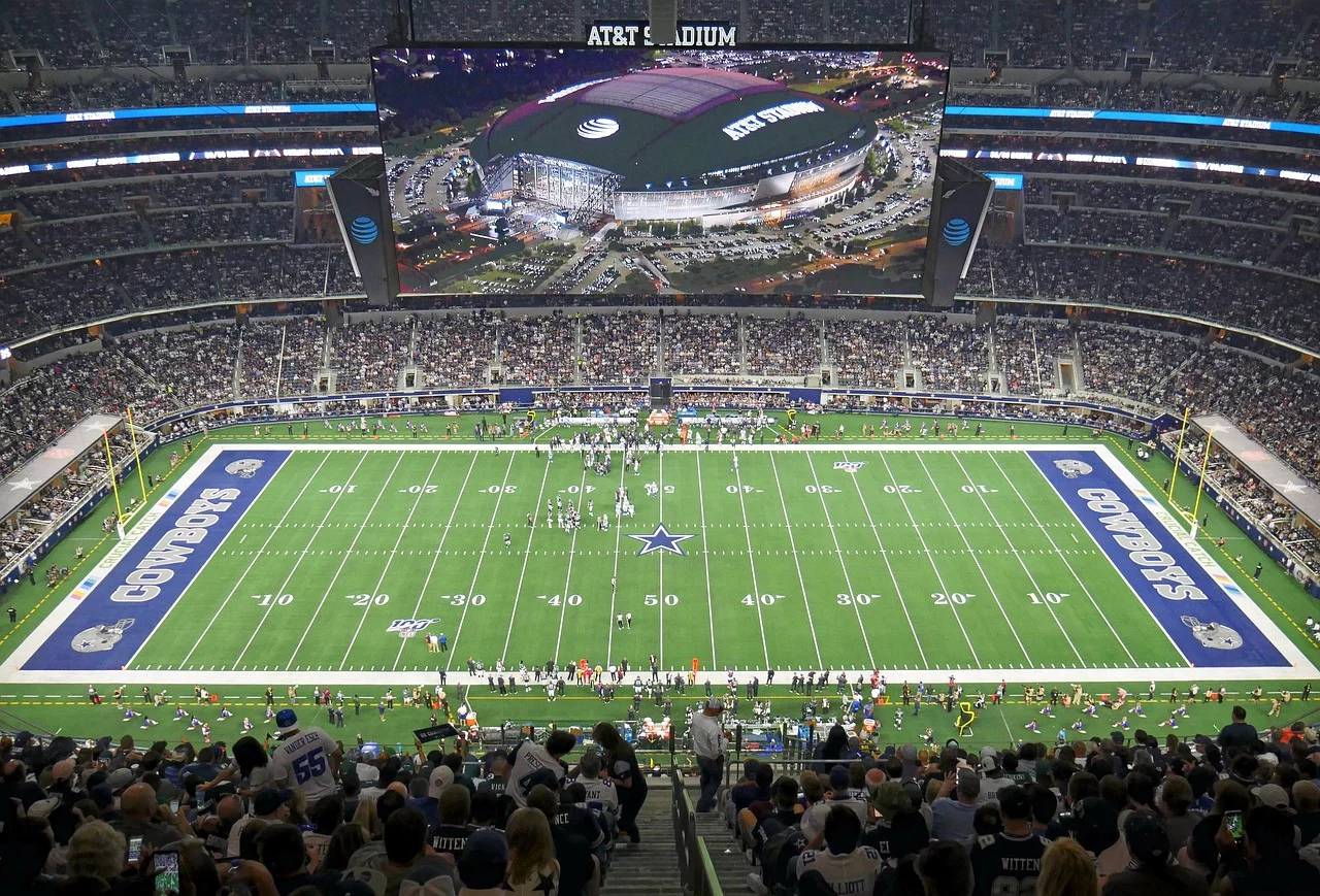 FootballR - NFL - Klayton Adams - Eine Weitwinkelansicht des AT&T Stadium während eines Footballspiels der Dallas Cowboys. Das Spielfeld, umgeben von einer großen Zuschauermenge, zeigt das Sternenlogo der Cowboys an der 50-Yard-Linie und in jeder Endzone steht „Cowboys“. Während die kommende Saison näher rückt, machen die Fans aufgeregt NFL-Wetten, während sie das Spiel auf einem großen Bildschirm an der Decke verfolgen. Diese Beschreibung wurde automatisch generiert.