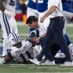 FootballR - NFL - Micah Parsons - Ein Footballspieler in weiß-blauer Uniform sitzt auf dem Feld und hält sein Bein. Auf seinem Trikot sind der Name „Parsons“ und die Nummer 11 zu sehen. Mehrere Teamkollegen und ein Trainer oder Sanitäter in weißem Hemd und blauer Hose stehen um ihn herum und bieten Micah Parsons Hilfe an. Diese Beschreibung wurde automatisch generiert.