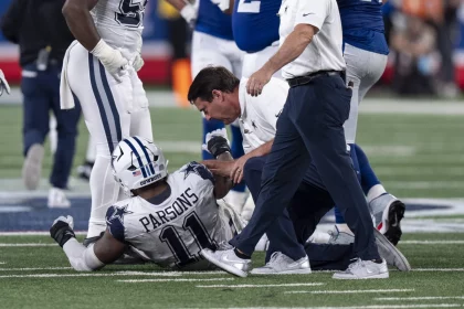 FootballR - NFL - Micah Parsons - Ein Footballspieler in weiß-blauer Uniform sitzt auf dem Feld und hält sein Bein. Auf seinem Trikot sind der Name „Parsons“ und die Nummer 11 zu sehen. Mehrere Teamkollegen und ein Trainer oder Sanitäter in weißem Hemd und blauer Hose stehen um ihn herum und bieten Micah Parsons Hilfe an. Diese Beschreibung wurde automatisch generiert.