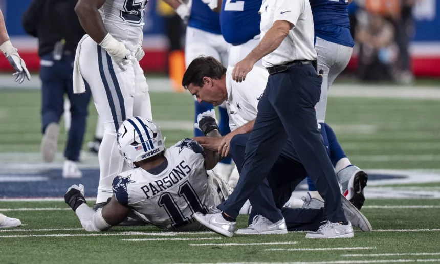 FootballR - NFL - Micah Parsons - Ein Footballspieler in weiß-blauer Uniform sitzt auf dem Feld und hält sein Bein. Auf seinem Trikot sind der Name „Parsons“ und die Nummer 11 zu sehen. Mehrere Teamkollegen und ein Trainer oder Sanitäter in weißem Hemd und blauer Hose stehen um ihn herum und bieten Micah Parsons Hilfe an. Diese Beschreibung wurde automatisch generiert.