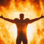 FootballR - NFL - Ein Fußballspieler im Trikot mit der Nummer 30 steht mit ausgestreckten Armen vor einem feurig orange-gelben Hintergrund. Das intensive Glühen und der Rauch erzeugen eine dramatische Szene und betonen die triumphale und kraftvolle Pose des Spielers. Diese Beschreibung wurde automatisch generiert.