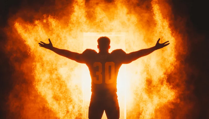 FootballR - NFL - Ein Fußballspieler im Trikot mit der Nummer 30 steht mit ausgestreckten Armen vor einem feurig orange-gelben Hintergrund. Das intensive Glühen und der Rauch erzeugen eine dramatische Szene und betonen die triumphale und kraftvolle Pose des Spielers. Diese Beschreibung wurde automatisch generiert.