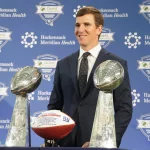 FootballR - NFL - Ein Mann im Anzug steht lächelnd hinter zwei silbernen Football-Trophäen und einem Football mit dem Logo der New York Giants. Im Hintergrund sind Logos von Hackensack Meridian Health und Quest Diagnostics Training Center zu sehen. Die Veranstaltung scheint ein formelles Medienereignis zu sein, an dem Eli Mannings Aufnahme in die Hall of Fame 2025 gefeiert wird. Diese Beschreibung wurde automatisch generiert.