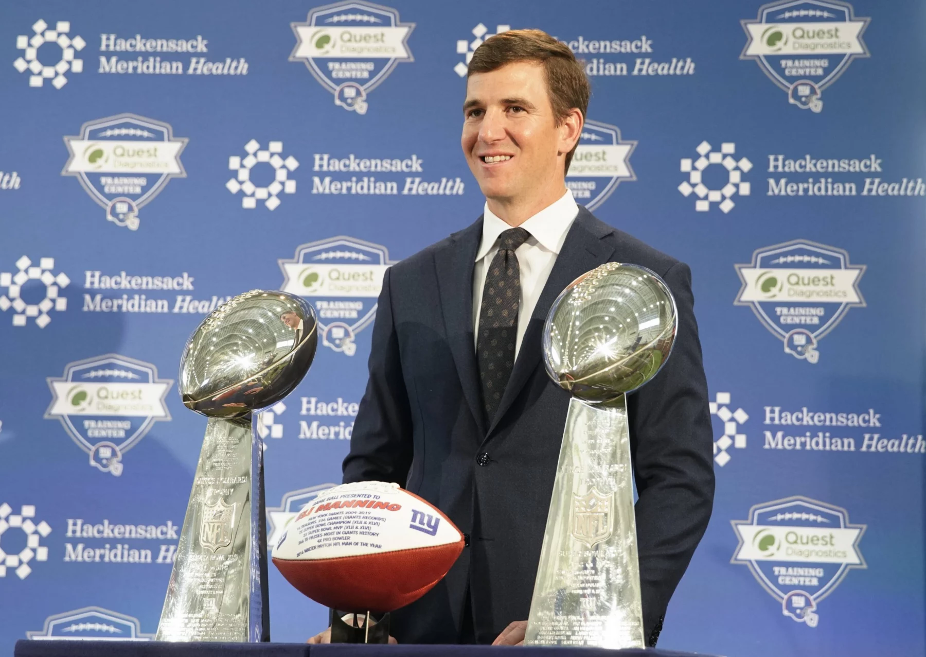 FootballR - NFL - Ein Mann im Anzug steht lächelnd hinter zwei silbernen Football-Trophäen und einem Football mit dem Logo der New York Giants. Im Hintergrund sind Logos von Hackensack Meridian Health und Quest Diagnostics Training Center zu sehen. Die Veranstaltung scheint ein formelles Medienereignis zu sein, an dem Eli Mannings Aufnahme in die Hall of Fame 2025 gefeiert wird. Diese Beschreibung wurde automatisch generiert.