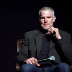 FootballR - NFL - Brett Favre - Ein Mann mit kurzen grauen Haaren und gestutztem Bart, der einen dunklen Blazer über einem schwarzen Hemd und Blue Jeans trägt, sitzt auf einem Stuhl vor einem dunklen Hintergrund. In seiner rechten Hand hält er ein Mikrofon, seine linke Hand ist teilweise zum Kinn erhoben und wirkt nachdenklich – er erinnert an die NFL-Legende Brett Favre. Diese Beschreibung wurde automatisch generiert.