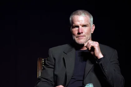 FootballR - NFL - Brett Favre - Ein Mann mit kurzen grauen Haaren und gestutztem Bart, der einen dunklen Blazer über einem schwarzen Hemd und Blue Jeans trägt, sitzt auf einem Stuhl vor einem dunklen Hintergrund. In seiner rechten Hand hält er ein Mikrofon, seine linke Hand ist teilweise zum Kinn erhoben und wirkt nachdenklich – er erinnert an die NFL-Legende Brett Favre. Diese Beschreibung wurde automatisch generiert.