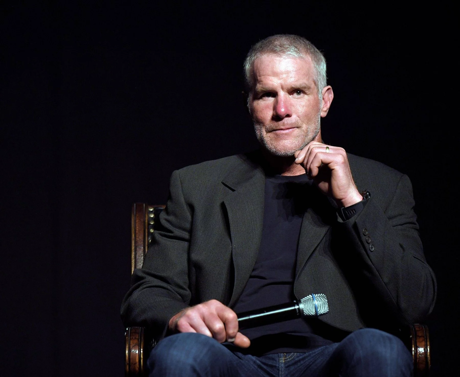 FootballR - NFL - Brett Favre - Ein Mann mit kurzen grauen Haaren und gestutztem Bart, der einen dunklen Blazer über einem schwarzen Hemd und Blue Jeans trägt, sitzt auf einem Stuhl vor einem dunklen Hintergrund. In seiner rechten Hand hält er ein Mikrofon, seine linke Hand ist teilweise zum Kinn erhoben und wirkt nachdenklich – er erinnert an die NFL-Legende Brett Favre. Diese Beschreibung wurde automatisch generiert.