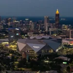 FootballR - NFL - Super Bowl LXII - Super Bowl 2028 - Luftaufnahme der Skyline von Atlanta in der Abenddämmerung mit beleuchteten Gebäuden. Im Vordergrund steht das Mercedes-Benz-Stadion, ein großes, modernes Gebäude mit einem einzigartigen eckigen Design, in dem der Super Bowl 2028 stattfinden soll. Im Hintergrund sind die Wolkenkratzer der Stadt zu sehen, darunter die in Gold beleuchtete Bank of America Plaza. Diese Beschreibung wurde automatisch generiert.