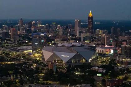 FootballR - NFL - Super Bowl LXII - Super Bowl 2028 - Luftaufnahme der Skyline von Atlanta in der Abenddämmerung mit beleuchteten Gebäuden. Im Vordergrund steht das Mercedes-Benz-Stadion, ein großes, modernes Gebäude mit einem einzigartigen eckigen Design, in dem der Super Bowl 2028 stattfinden soll. Im Hintergrund sind die Wolkenkratzer der Stadt zu sehen, darunter die in Gold beleuchtete Bank of America Plaza. Diese Beschreibung wurde automatisch generiert.