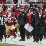 FootballR - RTL NFL - Christian McCaffrey, in rot-goldener Uniform, Nummer 23, läuft mit dem Ball an der Seitenlinie entlang. Er wird von einem Schiedsrichter und dem Trainerstab seines Teams, darunter Kyle Shanahan, verfolgt, die ihn aufmerksam beobachten. Im Hintergrund ist die Menge im Stadion zu sehen. Diese Beschreibung wurde automatisch generiert.