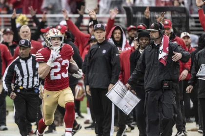 FootballR - RTL NFL - Christian McCaffrey, in rot-goldener Uniform, Nummer 23, läuft mit dem Ball an der Seitenlinie entlang. Er wird von einem Schiedsrichter und dem Trainerstab seines Teams, darunter Kyle Shanahan, verfolgt, die ihn aufmerksam beobachten. Im Hintergrund ist die Menge im Stadion zu sehen. Diese Beschreibung wurde automatisch generiert.