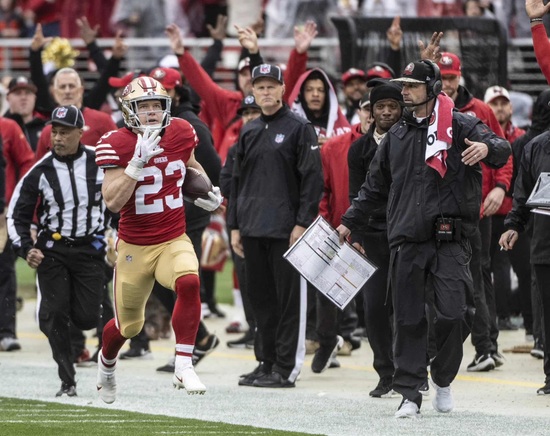 FootballR - RTL NFL - Christian McCaffrey, in rot-goldener Uniform, Nummer 23, läuft mit dem Ball an der Seitenlinie entlang. Er wird von einem Schiedsrichter und dem Trainerstab seines Teams, darunter Kyle Shanahan, verfolgt, die ihn aufmerksam beobachten. Im Hintergrund ist die Menge im Stadion zu sehen. Diese Beschreibung wurde automatisch generiert.