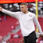 FootballR - NFL - Ein Footballspieler, möglicherweise Christian McCaffrey, trägt ein weißes Langarmshirt mit einem 49ers-Logo, schwarze Shorts und weiße Kopfhörer und streckt sein linkes Bein, während er auf seinem rechten Bein balanciert. Er streckt seinen linken Arm nach außen. Im Hintergrund ist ein leeres Stadion mit roten Sitzen und einem gelben Torpfosten zu sehen. Diese Beschreibung wurde automatisch generiert.