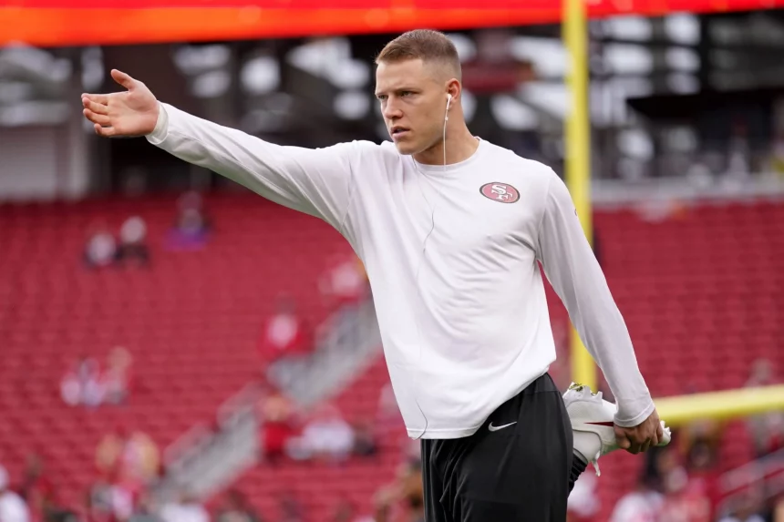 FootballR - NFL - Ein Footballspieler, möglicherweise Christian McCaffrey, trägt ein weißes Langarmshirt mit einem 49ers-Logo, schwarze Shorts und weiße Kopfhörer und streckt sein linkes Bein, während er auf seinem rechten Bein balanciert. Er streckt seinen linken Arm nach außen. Im Hintergrund ist ein leeres Stadion mit roten Sitzen und einem gelben Torpfosten zu sehen. Diese Beschreibung wurde automatisch generiert.