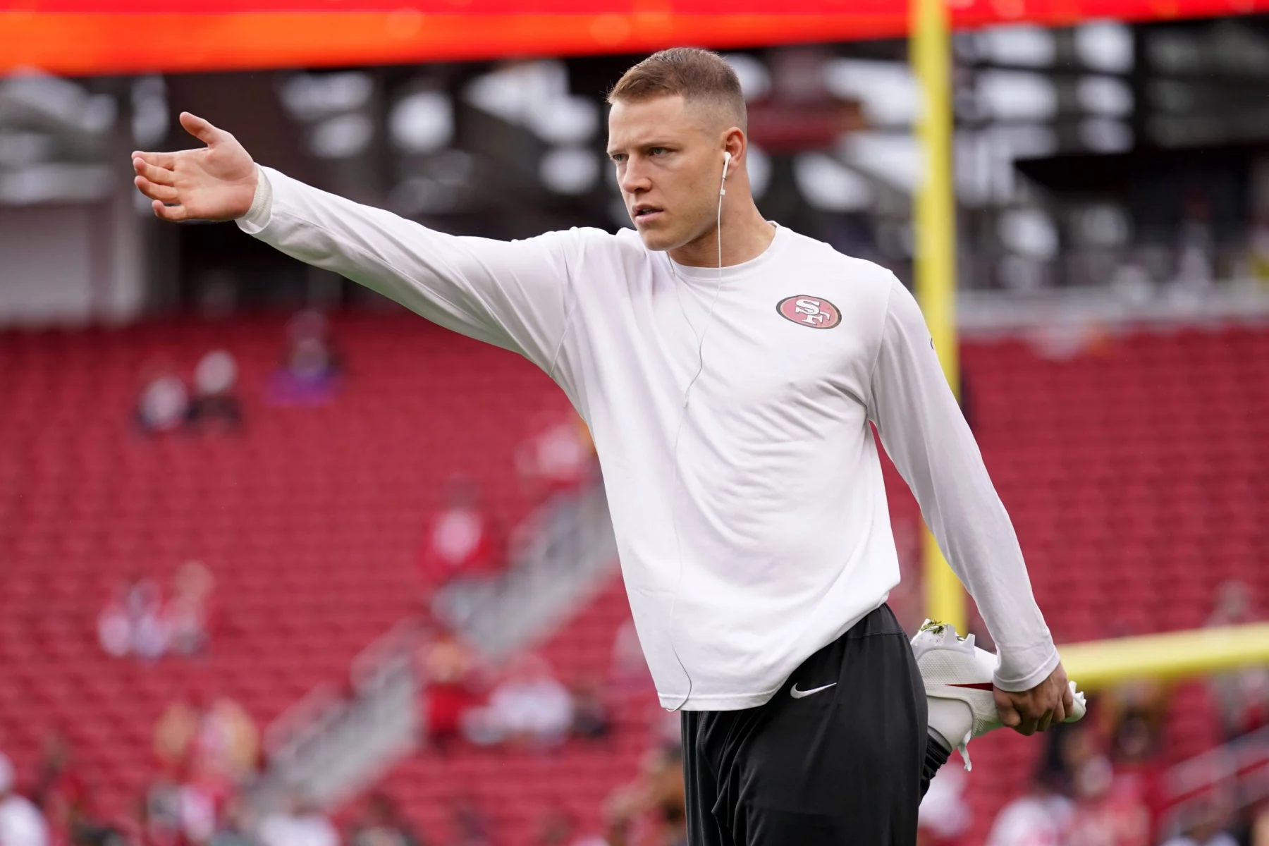 FootballR - NFL - Ein Footballspieler, möglicherweise Christian McCaffrey, trägt ein weißes Langarmshirt mit einem 49ers-Logo, schwarze Shorts und weiße Kopfhörer und streckt sein linkes Bein, während er auf seinem rechten Bein balanciert. Er streckt seinen linken Arm nach außen. Im Hintergrund ist ein leeres Stadion mit roten Sitzen und einem gelben Torpfosten zu sehen. Diese Beschreibung wurde automatisch generiert.