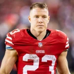FootballR - NFL - die meistgeklickten Artikel - Ein Footballspieler, möglicherweise Christian McCaffrey, trägt ein rotes Trikot mit der Nummer 23 und dem „49ers“-Logo und steht auf dem Spielfeld. Das Trikot sieht schmutzig aus, was darauf hindeutet, dass er erst vor kurzem gespielt hat. Der Gesichtsausdruck des Spielers ist konzentriert und im Hintergrund sind verschwommene Figuren und Stadionlichter zu sehen. Diese Beschreibung wurde automatisch generiert.