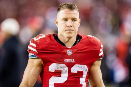 FootballR - NFL - die meistgeklickten Artikel - Ein Footballspieler, möglicherweise Christian McCaffrey, trägt ein rotes Trikot mit der Nummer 23 und dem „49ers“-Logo und steht auf dem Spielfeld. Das Trikot sieht schmutzig aus, was darauf hindeutet, dass er erst vor kurzem gespielt hat. Der Gesichtsausdruck des Spielers ist konzentriert und im Hintergrund sind verschwommene Figuren und Stadionlichter zu sehen. Diese Beschreibung wurde automatisch generiert.