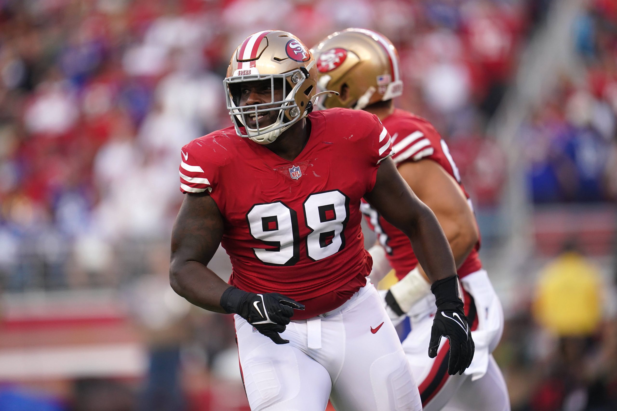 FootballR - NFL - Ein Footballspieler, der ein rotes San Francisco 49ers-Trikot mit der Nummer 98 und einen goldenen Helm trägt und als Javon Hargrave identifiziert wird, steht auf dem Spielfeld. Er ist in Bewegung, hinter ihm ist ein weiterer Spieler in einem weißen Trikot zu sehen. Im Hintergrund ist eine verschwommene Menschenmenge im Stadion zu sehen, darunter Zuschauer und ein Spieler in blau-weißer Uniform. Diese Beschreibung wurde automatisch generiert.