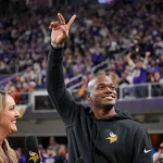 FootballR - NFL - Ein Mann in einem schwarzen Kapuzenpullover mit dem Logo der Minnesota Vikings winkt einer jubelnden Menge in einem Stadion zu. Er hält mit seiner rechten Hand ein Peace-Zeichen hoch. Der ehemalige Star-RB Adrian Peterson lächelt, während eine Frau mit langen blonden Haaren, die ein Mikrofon hält, in seiner Nähe steht. Fans in Lila jubeln wild. Diese Beschreibung wurde automatisch generiert.
