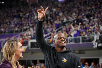 FootballR - NFL - Ein Mann in einem schwarzen Kapuzenpullover mit dem Logo der Minnesota Vikings winkt einer jubelnden Menge in einem Stadion zu. Er hält mit seiner rechten Hand ein Peace-Zeichen hoch. Der ehemalige Star-RB Adrian Peterson lächelt, während eine Frau mit langen blonden Haaren, die ein Mikrofon hält, in seiner Nähe steht. Fans in Lila jubeln wild. Diese Beschreibung wurde automatisch generiert.