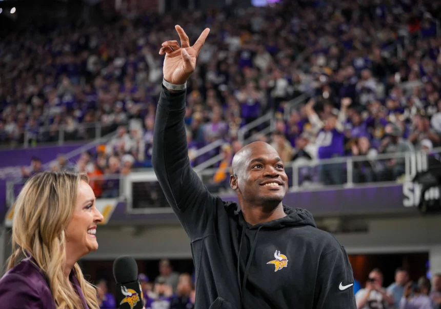 FootballR - NFL - Ein Mann in einem schwarzen Kapuzenpullover mit dem Logo der Minnesota Vikings winkt einer jubelnden Menge in einem Stadion zu. Er hält mit seiner rechten Hand ein Peace-Zeichen hoch. Der ehemalige Star-RB Adrian Peterson lächelt, während eine Frau mit langen blonden Haaren, die ein Mikrofon hält, in seiner Nähe steht. Fans in Lila jubeln wild. Diese Beschreibung wurde automatisch generiert.