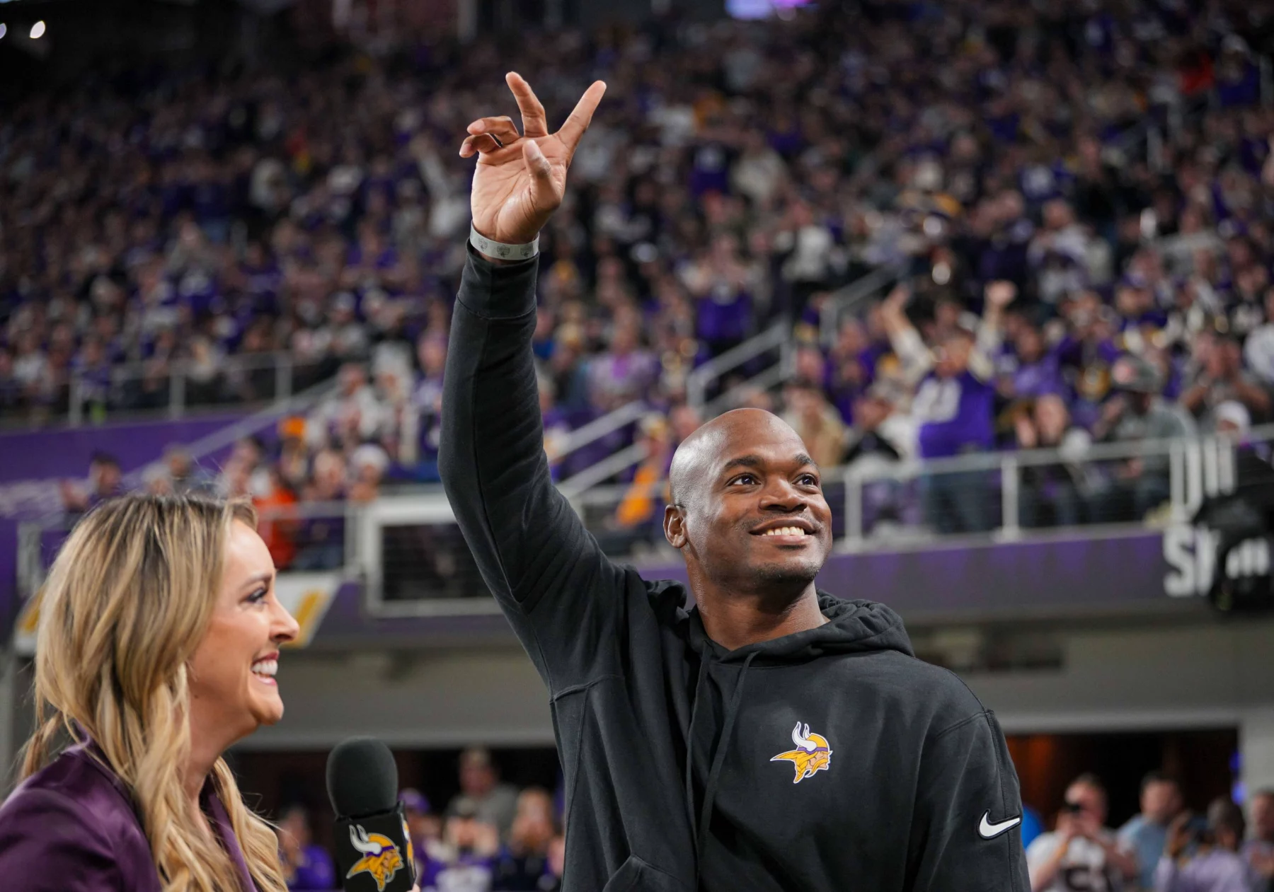 FootballR - NFL - Ein Mann in einem schwarzen Kapuzenpullover mit dem Logo der Minnesota Vikings winkt einer jubelnden Menge in einem Stadion zu. Er hält mit seiner rechten Hand ein Peace-Zeichen hoch. Der ehemalige Star-RB Adrian Peterson lächelt, während eine Frau mit langen blonden Haaren, die ein Mikrofon hält, in seiner Nähe steht. Fans in Lila jubeln wild. Diese Beschreibung wurde automatisch generiert.