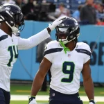 FootballR - NFL Bye Week - Zwei Spieler der Seattle Seahawks in weißen Trikots und Helmen stehen auf einem Footballfeld. Einer der Spieler, Kenneth Walker, lächelt, während sein Mundschutz an seiner Gesichtsmaske hängt. Er trägt die Nummer 9 und einen grünen Mundschutz. Der andere Spieler klopft spielerisch auf seinen Helm. Auf den Tribünen hinter ihnen stehen Fans und ein „SEE TO“-Schild. Diese Beschreibung wurde automatisch generiert.