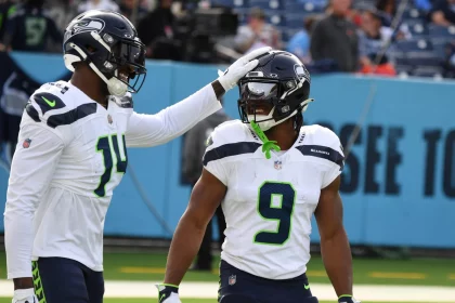 FootballR - NFL Bye Week - Zwei Spieler der Seattle Seahawks in weißen Trikots und Helmen stehen auf einem Footballfeld. Einer der Spieler, Kenneth Walker, lächelt, während sein Mundschutz an seiner Gesichtsmaske hängt. Er trägt die Nummer 9 und einen grünen Mundschutz. Der andere Spieler klopft spielerisch auf seinen Helm. Auf den Tribünen hinter ihnen stehen Fans und ein „SEE TO“-Schild. Diese Beschreibung wurde automatisch generiert.