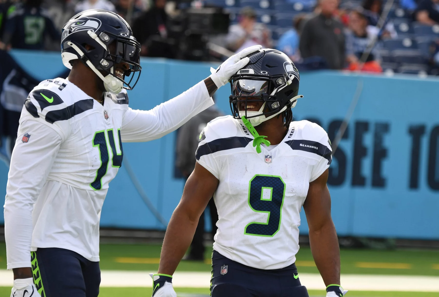 FootballR - NFL Bye Week - Zwei Spieler der Seattle Seahawks in weißen Trikots und Helmen stehen auf einem Footballfeld. Einer der Spieler, Kenneth Walker, lächelt, während sein Mundschutz an seiner Gesichtsmaske hängt. Er trägt die Nummer 9 und einen grünen Mundschutz. Der andere Spieler klopft spielerisch auf seinen Helm. Auf den Tribünen hinter ihnen stehen Fans und ein „SEE TO“-Schild. Diese Beschreibung wurde automatisch generiert.