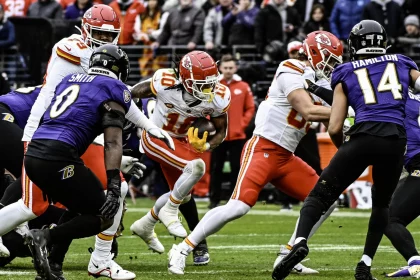 FootballR - NFL Roquan Smith - Im NFL-Saisonauftakt treffen Spieler der Kansas City Chiefs und der Baltimore Ravens auf dem Spielfeld aufeinander. Ein Chiefs-Spieler mit der Nummer 10 stürmt durch die Abwehr, während Ravens-Spieler Roquan Smith und ein weiterer Spieler mit der Nummer 14 versuchen, ihn zu blockieren. Im Hintergrund dieses spannenden Spiels zwischen den Ravens und den Chiefs jubeln begeisterte Fans. Diese Beschreibung wurde automatisch generiert.