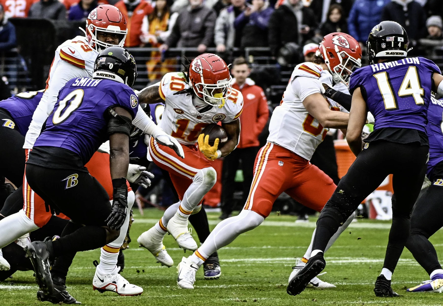 FootballR - NFL Roquan Smith - Im NFL-Saisonauftakt treffen Spieler der Kansas City Chiefs und der Baltimore Ravens auf dem Spielfeld aufeinander. Ein Chiefs-Spieler mit der Nummer 10 stürmt durch die Abwehr, während Ravens-Spieler Roquan Smith und ein weiterer Spieler mit der Nummer 14 versuchen, ihn zu blockieren. Im Hintergrund dieses spannenden Spiels zwischen den Ravens und den Chiefs jubeln begeisterte Fans. Diese Beschreibung wurde automatisch generiert.