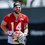 FootballR - NFL - Ricky Pearsall - Ein Footballspieler, der ein rotes Trainingstrikot mit der Nummer 14, ein weißes Unterhemd und ein Stirnband trägt, grinst und hält einen Helm der San Francisco 49ers. Das Trainingsfeld ist im Hintergrund verschwommen, und neben ihm ist ein weiterer WR der 49ers in einem ähnlichen roten Trikot teilweise sichtbar. Diese Beschreibung wurde automatisch generiert.