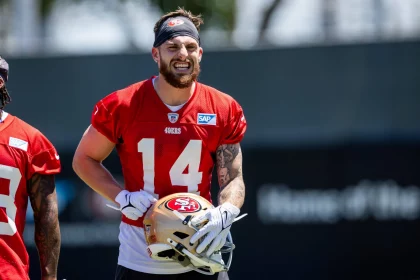FootballR - NFL - Ricky Pearsall - Ein Footballspieler, der ein rotes Trainingstrikot mit der Nummer 14, ein weißes Unterhemd und ein Stirnband trägt, grinst und hält einen Helm der San Francisco 49ers. Das Trainingsfeld ist im Hintergrund verschwommen, und neben ihm ist ein weiterer WR der 49ers in einem ähnlichen roten Trikot teilweise sichtbar. Diese Beschreibung wurde automatisch generiert.