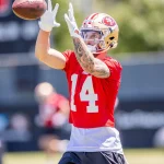 FootballR - NFL - Ricky Pearsall, der ein rotes Trikot mit der Nummer 14 und einen Helm der San Francisco 49ers trägt, streckt beim Training beide Hände aus, um einen Football zu fangen. Im Hintergrund ist ein verschwommenes Außenfeld mit anderen Spielern und einer Anzeigetafel zu sehen. Er hat Tätowierungen auf seinem rechten Arm. Diese Beschreibung wurde automatisch generiert.