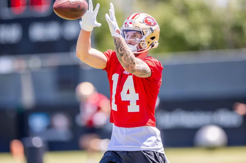 FootballR - NFL - Ricky Pearsall, der ein rotes Trikot mit der Nummer 14 und einen Helm der San Francisco 49ers trägt, streckt beim Training beide Hände aus, um einen Football zu fangen. Im Hintergrund ist ein verschwommenes Außenfeld mit anderen Spielern und einer Anzeigetafel zu sehen. Er hat Tätowierungen auf seinem rechten Arm. Diese Beschreibung wurde automatisch generiert.