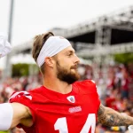 FootballR - NFL - Ein Footballspieler mit der Nummer 14 auf seinem rot-weißen 49ers-Trikot wirft ein weißes Handtuch in die Menge. Ricky Pearsall trägt ein weißes Stirnband und ein Band um seinen linken Ellbogen. Im Hintergrund sieht man ein Publikum, das von Tribünen aus zusieht, einige in roter Kleidung, und eine große Außenkonstruktion. Diese Beschreibung wurde automatisch generiert.