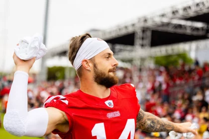 FootballR - NFL - Ein Footballspieler mit der Nummer 14 auf seinem rot-weißen 49ers-Trikot wirft ein weißes Handtuch in die Menge. Ricky Pearsall trägt ein weißes Stirnband und ein Band um seinen linken Ellbogen. Im Hintergrund sieht man ein Publikum, das von Tribünen aus zusieht, einige in roter Kleidung, und eine große Außenkonstruktion. Diese Beschreibung wurde automatisch generiert.