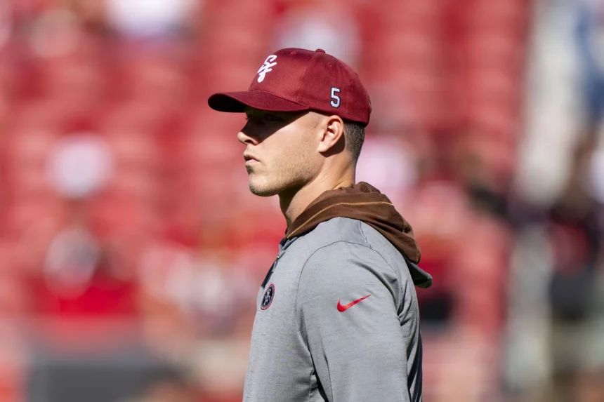 FootballR - NFL -Christian McCaffrey - Ein Mann mit einer roten Kappe und einem grauen Kapuzenpullover steht da. Auf der Kappe sind das 49ers-Logo und die Nummer 5 an der Seite zu sehen. Im Hintergrund ist ein verschwommenes Stadion mit roten Sitzen zu sehen, wahrscheinlich während des Saisonauftakts. Er scheint aufmerksam auf die linke Seite des Bildes zu blicken. Die Beleuchtung ist hell und deutet auf einen sonnigen Tag hin. Diese Beschreibung wurde automatisch generiert.