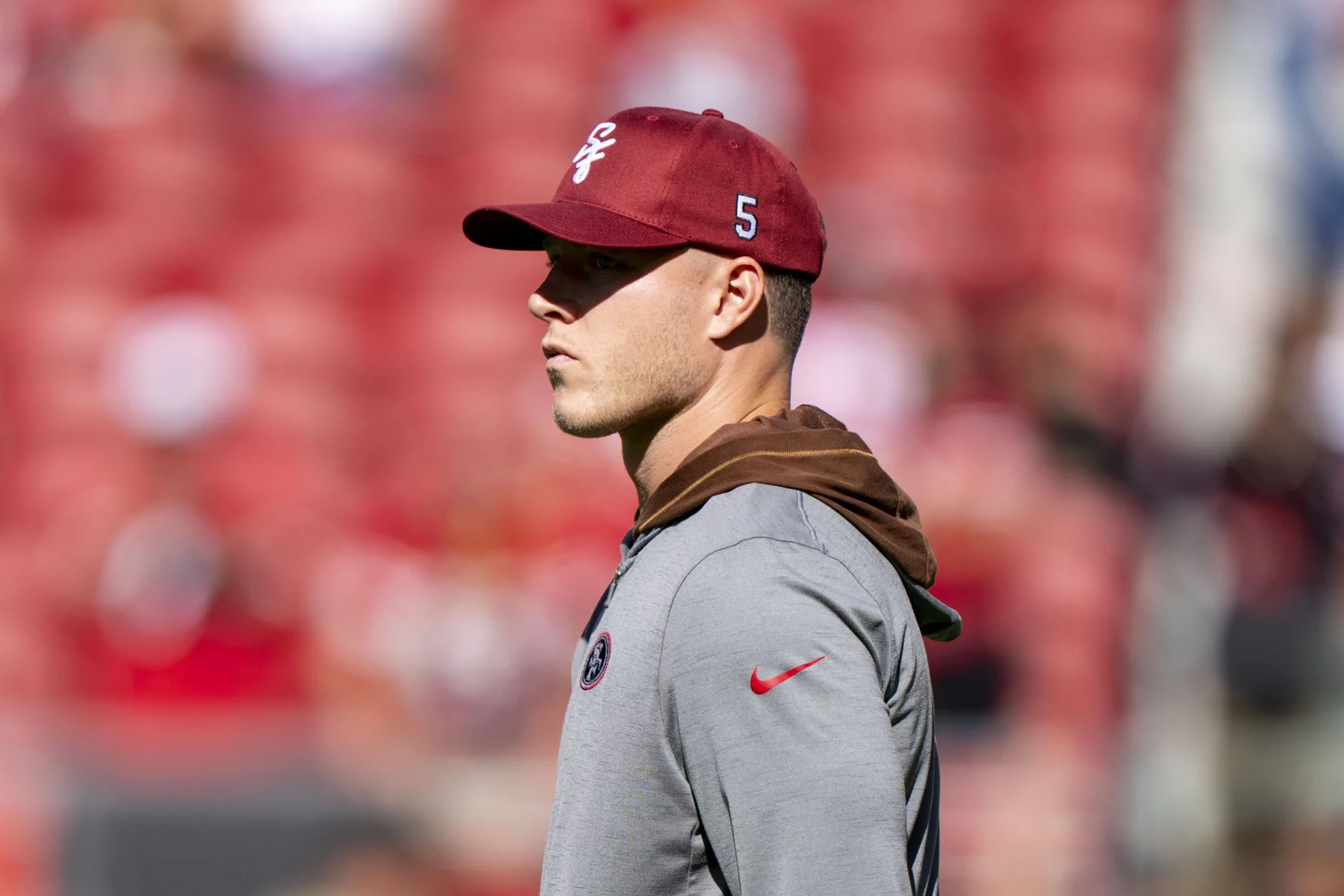 FootballR - NFL -Christian McCaffrey - Ein Mann mit einer roten Kappe und einem grauen Kapuzenpullover steht da. Auf der Kappe sind das 49ers-Logo und die Nummer 5 an der Seite zu sehen. Im Hintergrund ist ein verschwommenes Stadion mit roten Sitzen zu sehen, wahrscheinlich während des Saisonauftakts. Er scheint aufmerksam auf die linke Seite des Bildes zu blicken. Die Beleuchtung ist hell und deutet auf einen sonnigen Tag hin. Diese Beschreibung wurde automatisch generiert.