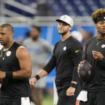 FootballR - NFL - Drei Footballspieler , darunter Russell Wilson, der Pittsburgh Steelers stehen auf dem Feld, alle tragen schwarze ärmellose Trikots und Stirnbänder. Einer hält einen Football in der Hand, während sie nach vorne blicken, was an Justin Fields' intensive Vorbereitung erinnert. Im Hintergrund ist ein Hallenstadion mit blauen Sitzen und verschwommenen Figuren zu sehen. Diese Beschreibung wurde automatisch generiert.