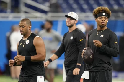 FootballR - NFL - Drei Footballspieler , darunter Russell Wilson, der Pittsburgh Steelers stehen auf dem Feld, alle tragen schwarze ärmellose Trikots und Stirnbänder. Einer hält einen Football in der Hand, während sie nach vorne blicken, was an Justin Fields' intensive Vorbereitung erinnert. Im Hintergrund ist ein Hallenstadion mit blauen Sitzen und verschwommenen Figuren zu sehen. Diese Beschreibung wurde automatisch generiert.