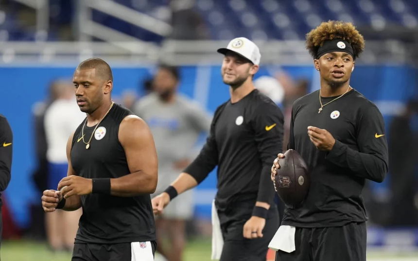 FootballR - NFL - Drei Footballspieler , darunter Russell Wilson, der Pittsburgh Steelers stehen auf dem Feld, alle tragen schwarze ärmellose Trikots und Stirnbänder. Einer hält einen Football in der Hand, während sie nach vorne blicken, was an Justin Fields' intensive Vorbereitung erinnert. Im Hintergrund ist ein Hallenstadion mit blauen Sitzen und verschwommenen Figuren zu sehen. Diese Beschreibung wurde automatisch generiert.